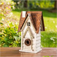 a white birdhouse with a brown roof sitting on a wooden table in front of some bushes