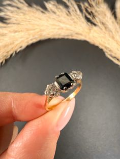 a hand holding an engagement ring with three stones on it and some dry grass in the background