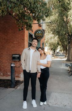 a man and woman standing in front of a brick building with trees on the sidewalk