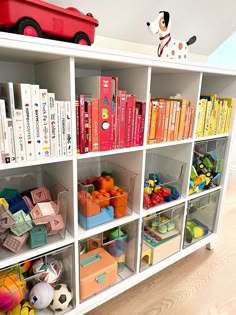 a white bookcase filled with lots of books next to a red toy truck on top of a hard wood floor