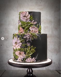 a three tiered cake decorated with flowers and greenery on a black stand in front of a concrete wall