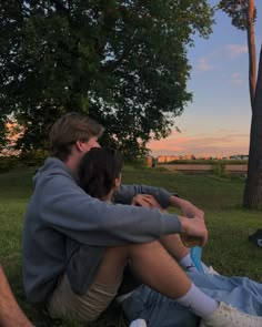 two people sitting on the grass in front of a tree with their arms around each other