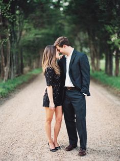 a man and woman standing in the middle of a dirt road with trees behind them