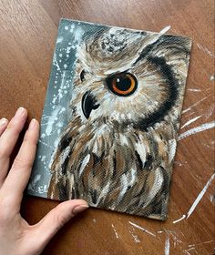 an owl painting being displayed on a wooden table with someone's hand holding it