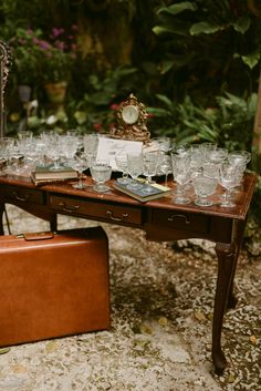 a table with many glasses on it and a suitcase sitting next to it in front of some plants