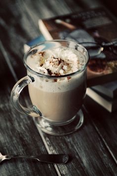 a cup of hot chocolate with whipped cream on top next to a book and spoon