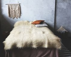 a bed with a white fur blanket and pillows on it's headboard, next to a gray wall