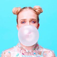 a woman blowing a bubble into her face with blue eyes and blonde hair, against a light blue background