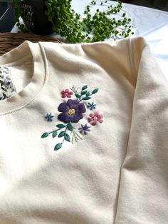 a white sweater with purple flowers embroidered on the front and side, sitting next to a plant