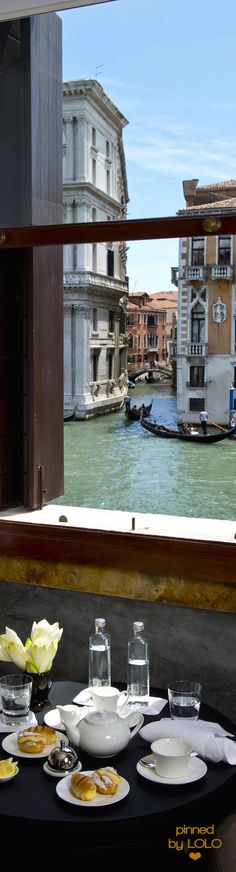 the table is set with food and water in front of an open window that overlooks a canal