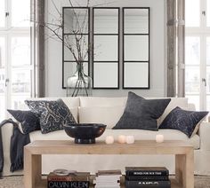 a living room with white couches and pillows on top of the coffee table in front of two windows