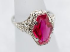 This fantastic Art Deco era ring is iconic of the early 1900s, with a perfectly preserved synthetic ruby gemstone set at its center. In the Victorian era, transportation was far more expensive than it is now. Rubies had to be brought from Burma, and Emeralds from South America. This cost translated into the price of the stones, making them far too expensive for the average consumer. In response, jewelers found interesting ways to make natural stones go a little farther. Synthetics were created: stones faceted from crystals grown in laboratories. These remained popular right through the Art Deco era. Metal: 18K White GoldGem: Synthetic Ruby Gem Measurements: 12.0 x 8.1 mm, Fancy CutRing Size: 4Marks: “18” Stamped on the inside band Vintage Rings With Octagon Center Stone, Vintage Octagon Ring With Center Stone, Fine Jewelry Ruby Signet Ring For Formal Occasions, Formal Ruby Signet Ring In Fine Jewelry Style, Ruby Signet Ring Fine Jewelry For Formal Events, Ruby Signet Ring For Formal Occasions, Vintage Hallmarked White Gold Ruby Ring, Hallmarked Vintage White Gold Ruby Ring, Formal Ruby Ring