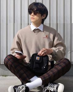 a young man sitting on the ground with his cell phone in his hand and wearing plaid pants