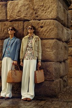 two women standing next to each other holding purses and bags in front of a stone wall