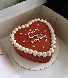 a red heart shaped cake with white frosting and hearts on the top is sitting in a box