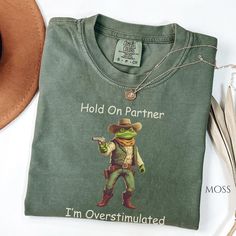 Hold on Partner, I'm Overstimulated Funny Frog Shirt, Cowboy Frog Tee, Unisex Ironic Shirt, Weirdcore Shirt. DETAILS .: Comfort Colors® Unisex Garment-dyed t-shirt. .: Made 100% with ring-spun cotton.  .: The soft-washed, garment-dyed fabric brings extra coziness to your wardrobe while the      relaxed fit makes it an excellent daily choice.  .: The double-needle stitching throughout the tee makes it highly durable while the lack     of side-seams helps the shirt retain its tubular shape. .: Direct To Garment Print, which means the dye does not sit on top like vinyl but is     embedded into the fabric. SIZING .: Please refer to our size chart for specifications. This shirt is designed to have a relaxed     fit. For a more fitted look, consider ordering a size down. For a more relaxed or Ironic Tshirt, Ironic Tshirts, Frog Meme, Funny Frogs, Iron Shirt, Gen Z, Comfort Color, Look Plus, Oversized Shirt