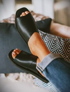 Close-up of a person's feet wearing Stormi Black Slide Sandals from Kay Kay Fashion. Their ankles are crossed and resting on a cushioned surface, showcasing women's flat shoes that are both comfortable and true to size. The person is wearing blue rolled-up jeans, with patterned cushions and soft furnishings in the background. Black Slides Women, Black Slide Sandals, Stylish Black Women, Black Work Shoes, Work Shoe, Chic Sandals, Black Slides, Latina Fashion, 2024 Style