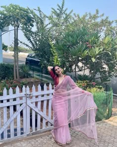 a woman in a pink sari standing next to a white fence with trees and bushes behind her