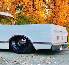 an old white car is parked in front of some trees with orange leaves on the ground