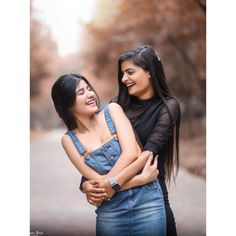 two women standing next to each other in front of trees and dirt road with their arms around each other