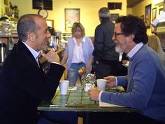 two men sitting at a table talking to each other in a restaurant with people standing around