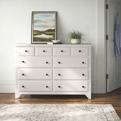 a white dresser sitting on top of a wooden floor next to a painting and potted plant