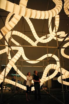 two people standing in front of a sculpture made out of white paper tubes and wires