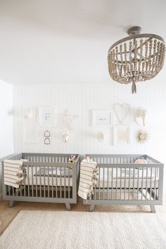 two baby cribs in a white room with pictures on the wall and a chandelier