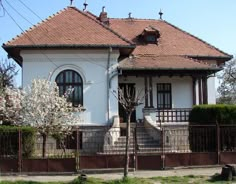 a white house with red tiled roof next to a fence and trees in front of it