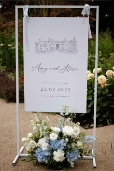 a sign that is sitting on the ground with flowers in front of it and an arrangement of blue and white flowers behind it