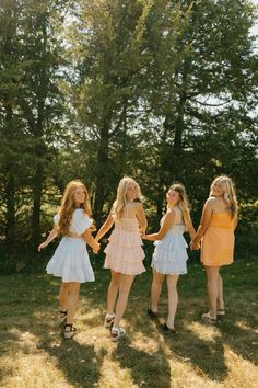 three girls in dresses are holding hands and walking through the grass with trees behind them