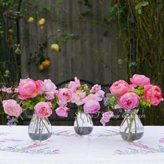 three vases filled with pink flowers sitting on top of a white tablecloth covered table