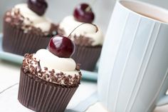 three cupcakes with chocolate frosting and cherries on top, next to a coffee mug