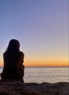 a woman sitting on top of a rock next to the ocean watching the sun go down