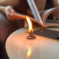 a woman reading a book while sitting on a chair with a lit candle in front of her