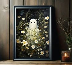 a framed painting of a ghost surrounded by daisies on a wooden table next to a candle