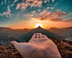 a person's hand on top of a mountain with the sun setting in the background