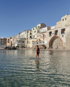 a woman wading in the water near some buildings and people walking on the beach