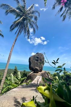 a large rock sitting on top of a lush green hillside next to the ocean and palm trees