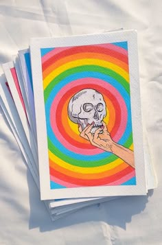 a hand holding a skull in front of a rainbow colored spiral background with white paper