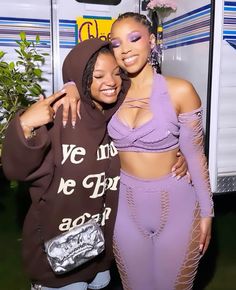 two women standing next to each other in front of a bus wearing purple outfits and matching makeup