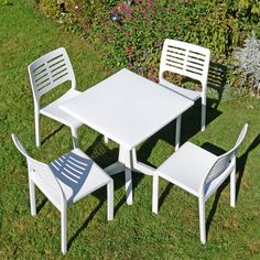 three white plastic chairs and a table in the middle of some grass with flowers behind it