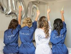 three bridesmaids in matching robes hold up their champagne flutes as they sit on a bed