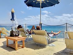 two people lounging on lounge chairs under an umbrella at the beach with blue water in the background