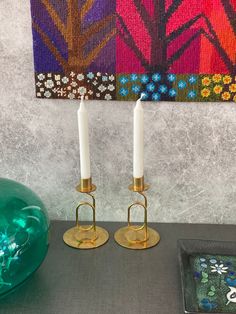 two candles are sitting on a table next to a green vase and decorative wall hanging