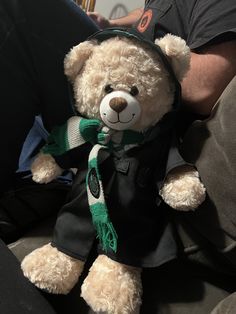 a brown teddy bear wearing a green and white scarf on it's lap with a man in the background