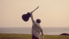 a man with a guitar in his hand on the grass by the ocean is looking at the sky