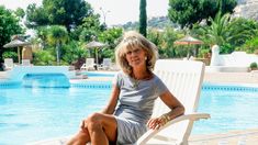 a woman sitting on a white chair next to a swimming pool with palm trees in the background