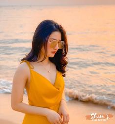 a woman in a yellow dress standing on the beach at sunset with her hands behind her back