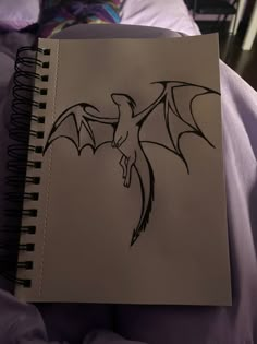 a drawing of a dragon on a notebook in bed with purple sheets and pillows behind it